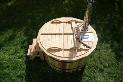 Round wooden barrel hot tub with lid, steps, and a metal chimney on a grassy lawn.